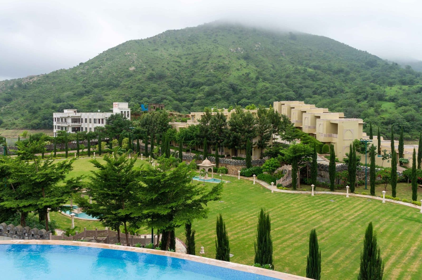A resort spread across a green hillside: sandstone wings, lawns lined with cypress trees and a pool in front.