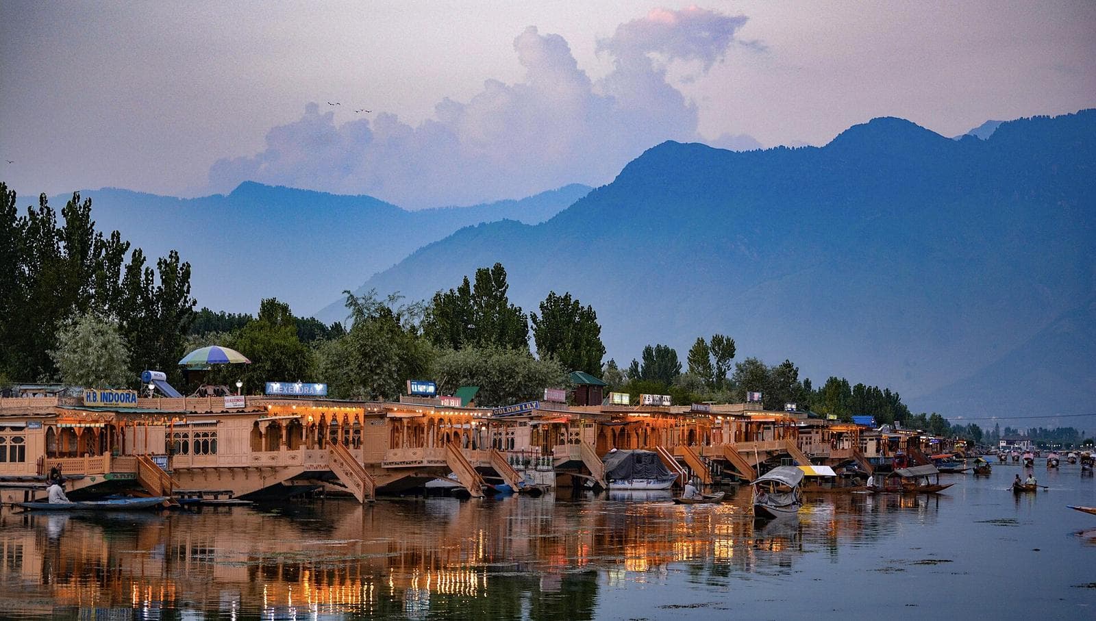 A row of carved wooden houseboats moored along Dal Lake, with mountains behind.