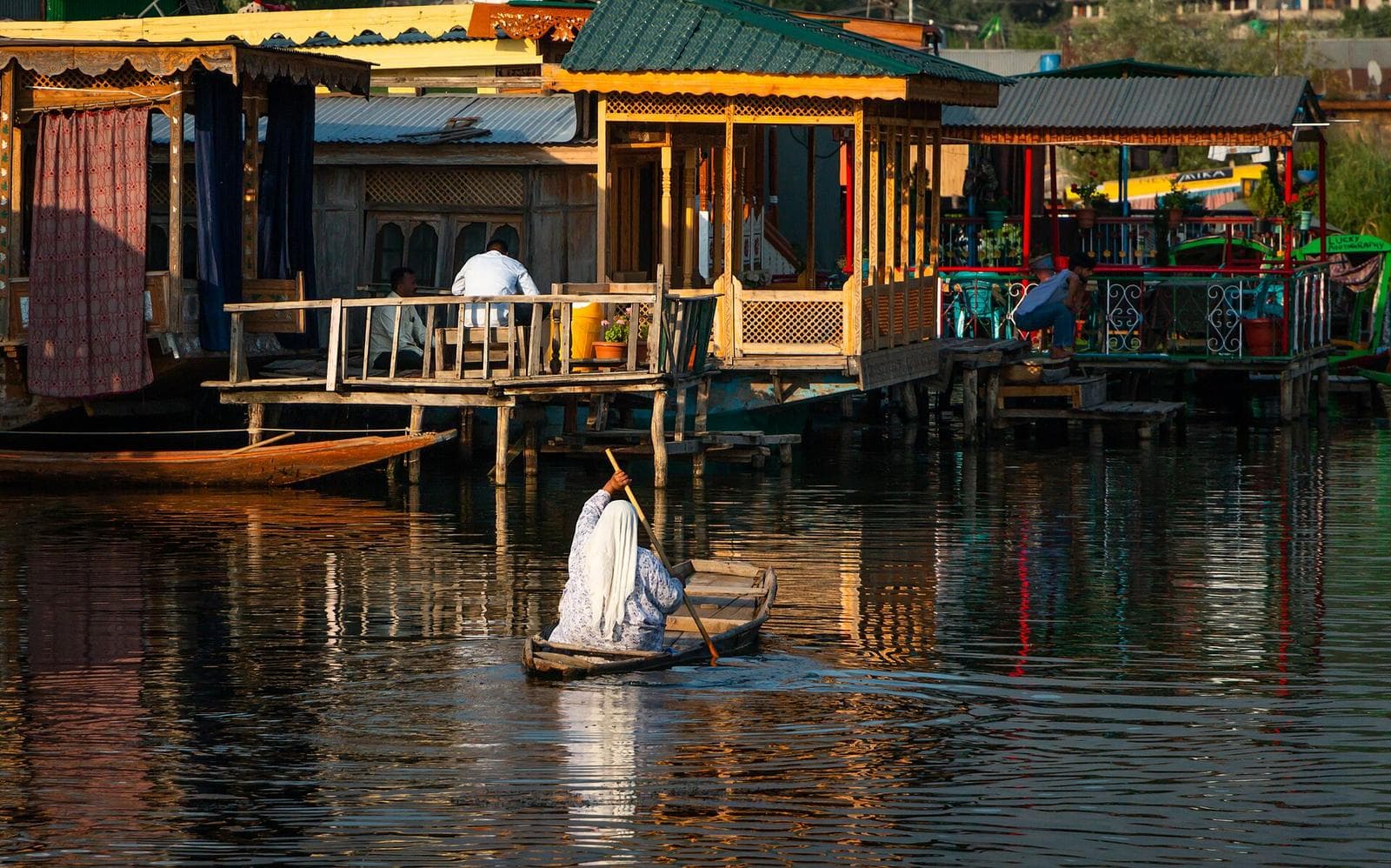 A woman rowing a shikara past the carved wooden balconies of a houseboat on Dal Lake.