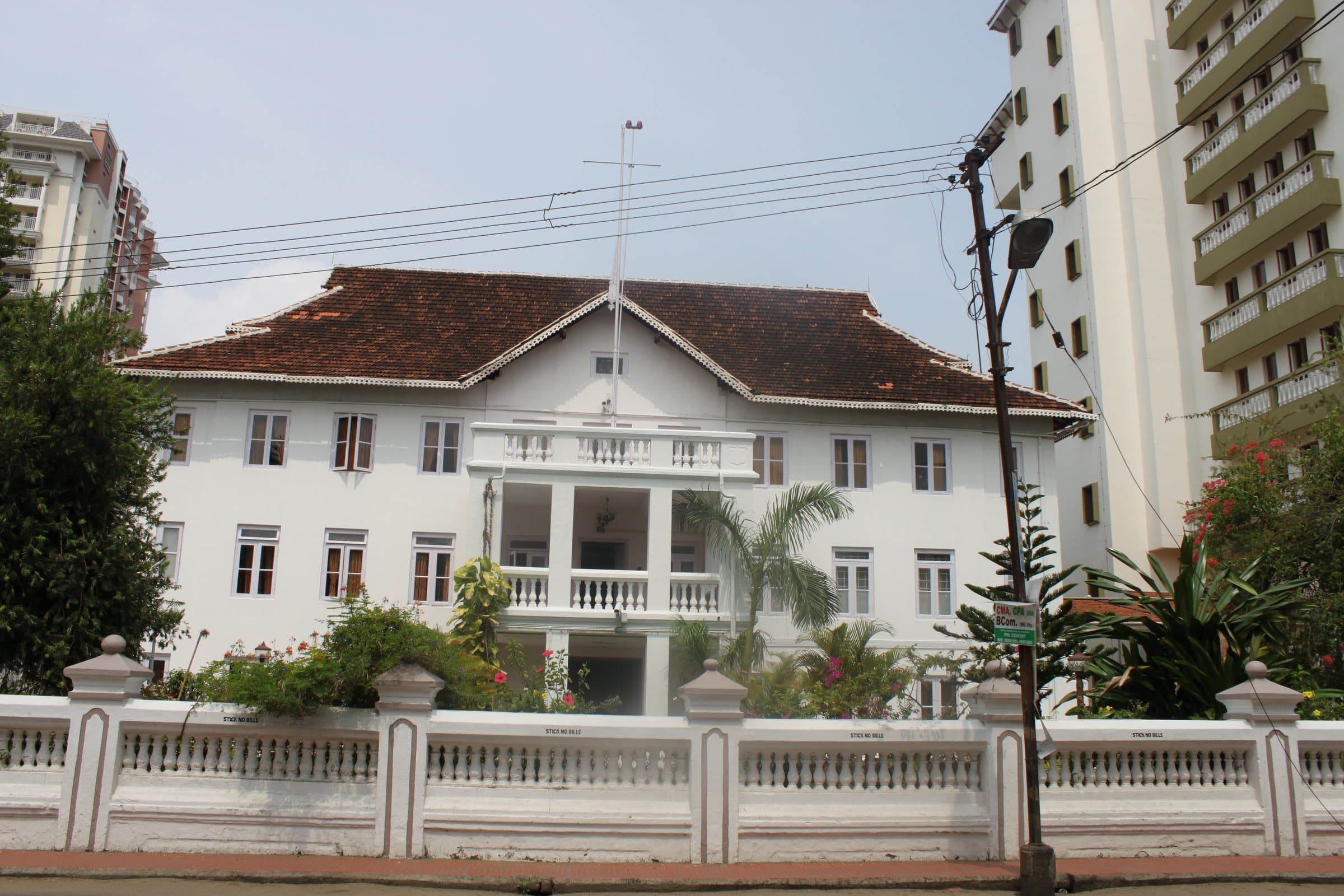 A white two-storey colonial building with a red tiled roof and a pillared balcony at its centre, behind a white balustrade wall, with palms in front and apartment blocks on either side.