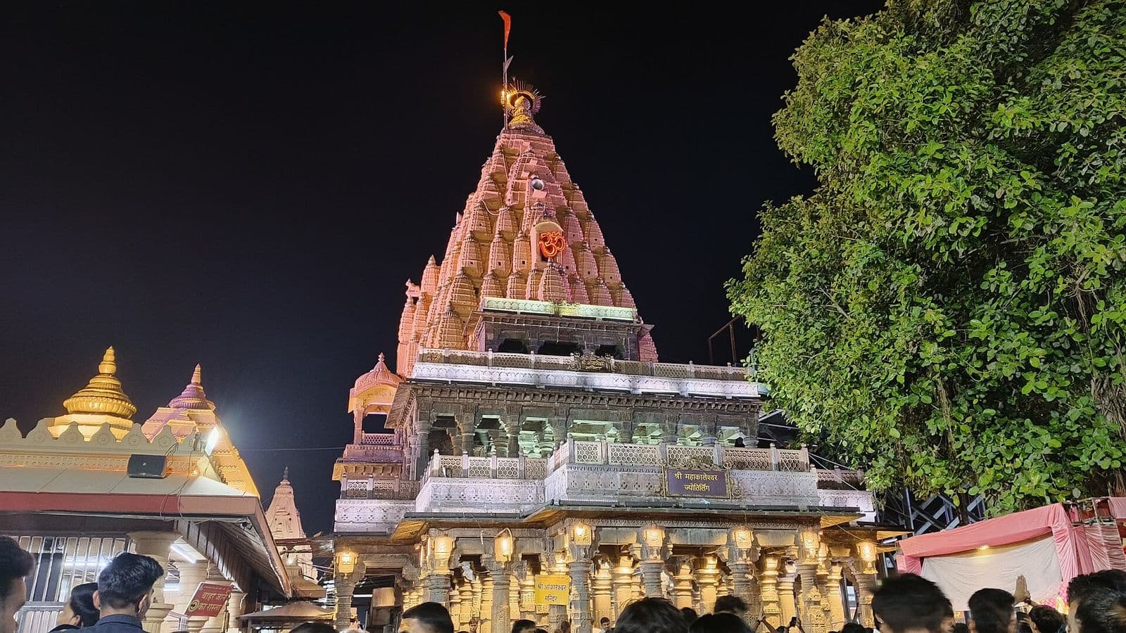 The carved spire of the Mahakaleshwar temple lit up at night, with devotees in the courtyard below.