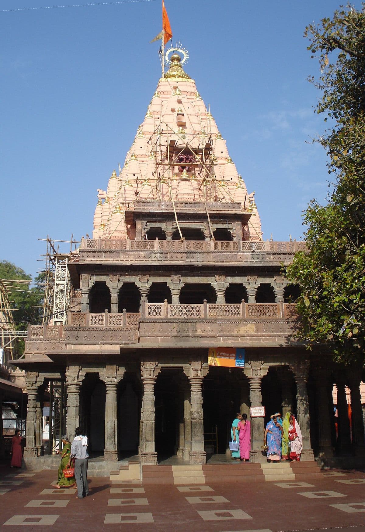 The tall carved spire of the Mahakaleshwar temple in Ujjain under a clear blue sky, above its pillared hall.