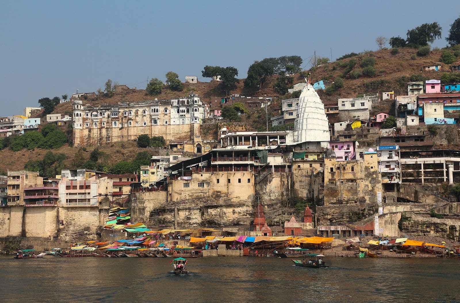 The white spire of the Omkareshwar temple rising from the town on a rocky island in the Narmada, with boats on the river.