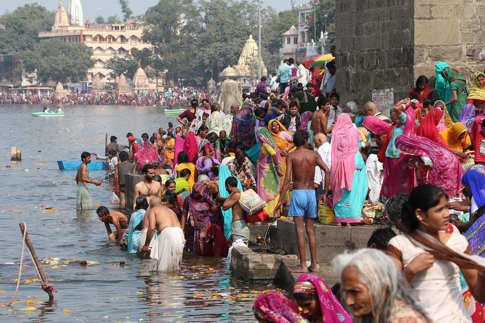 Pilgrims in bright saris bathing in the Shipra at Ram Ghat, with temples along the far bank.