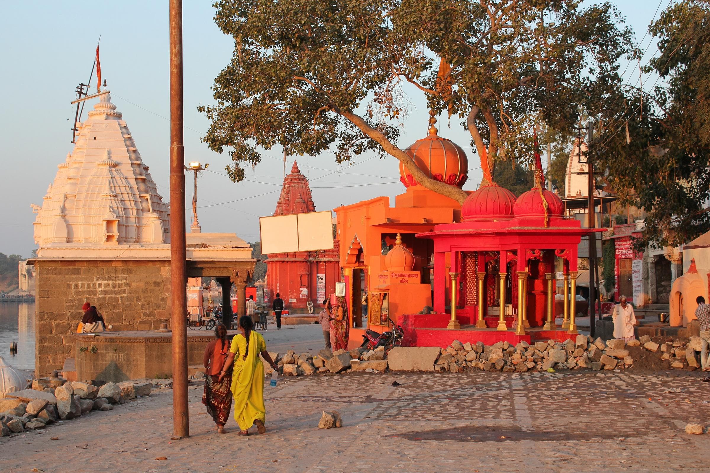 Small white and orange-red temples on Ram Ghat in Ujjain, lit by the late-afternoon sun, with two women walking past.