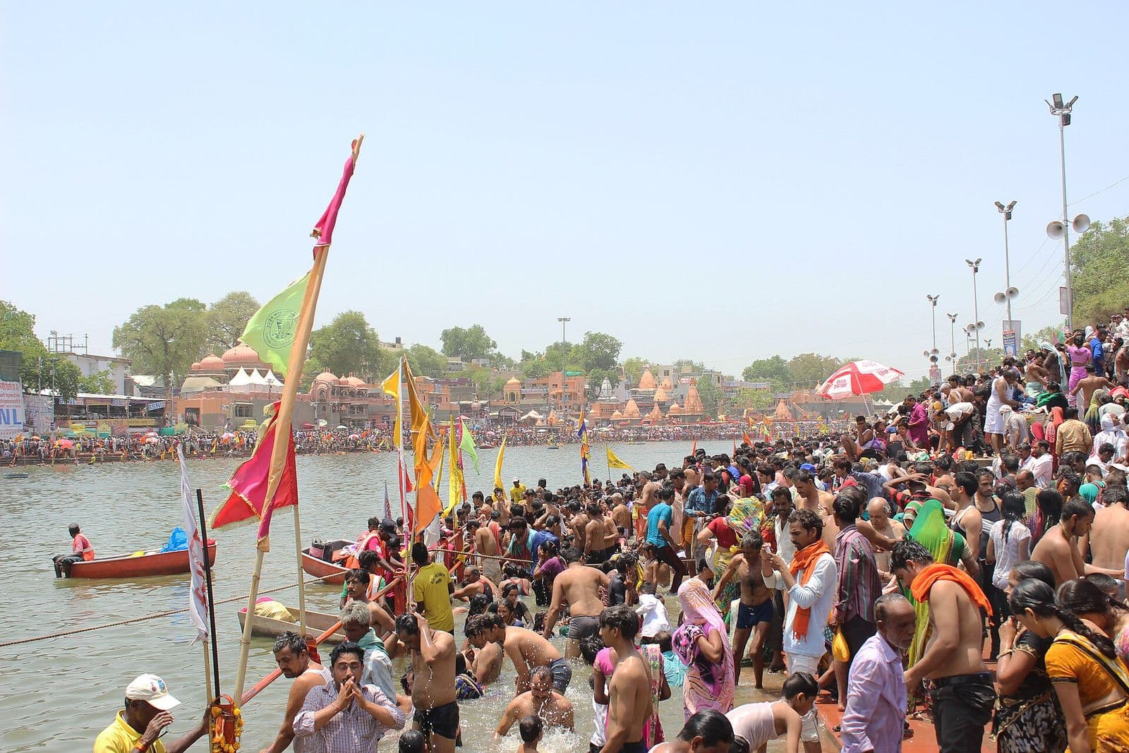 Crowds of pilgrims bathing in the Shipra during Simhastha 2016, with saffron and green flags on the ghat and boats on the river.